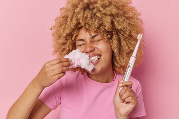 Glad young woman with curly bushy hair has sweet tooth eats candy floss going to clean teeth with electric brush dressed in casual t shirt isolated over pink background. Food bad for your teeth