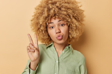 Pretty young European woman with curly bushy hair keeps lips folded shows victory v sign wears...