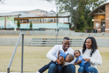 A beautiful African-American mom and dad sitting on steps outdoors and tickeling their two sons and everyone is laughing