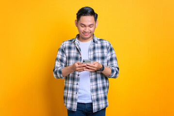 Smiling young Asian man in plaid shirt using mobile phone, reading good news isolated on yellow background