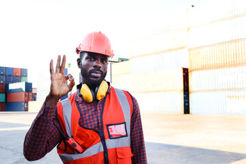 Portrait of African American young engineer worker man wearing safety bright neon red color vest and helmet, giving OK sign hand at logistic shipping cargo container yard.