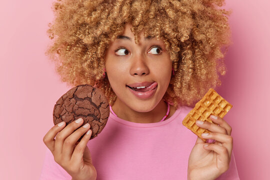 Photo of lovely female model with curly hair licks lips holds appetizing cookie and waffle enjoys snack time has sweet tooth wears casual t shirt isolated over pink background. Unhealthy food concept