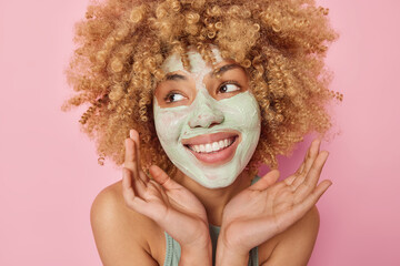 Headshot of cheerful curly woman applies beauty nourishing mask on face keeps palms over face looks away with glad expression smiles toothily stands bare shoulders isolated over pink background