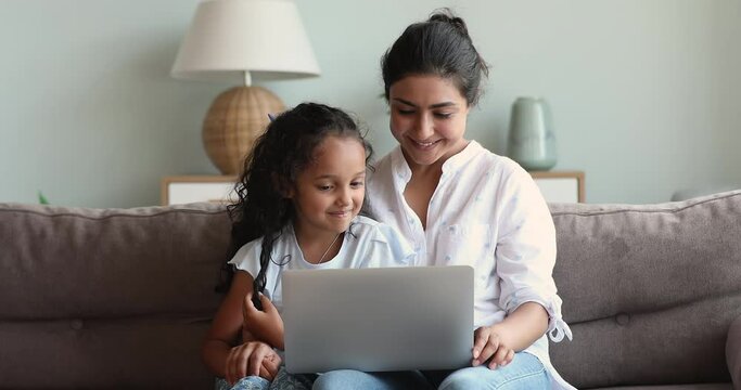 Adorable little girl of Indian ethnicity rest on sofa with single mother engaged in online web shopping with parent using laptop pc. Bonding mum gen z daughter preschooler talk surf sites on computer