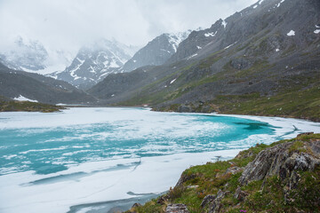 Obraz premium Atmospheric mountain landscape with frozen alpine lake during snowfall. Awesome cold scenery with snowfall in high mountain valley with icy mountain lake on background of snow mountains in low clouds.