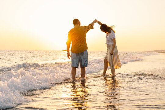 Young romantic couple dancing turning around by sea. Seascape at sunset with beautiful sky. Romantic couple on the beach at golden sunset. Lover couple having fun on beach.