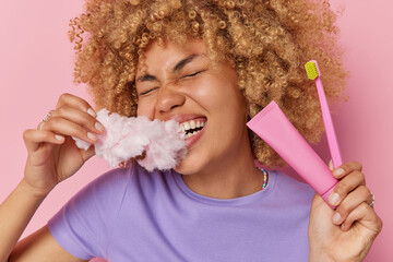 Cheerful curly haired woman affords herself eating high calorie harmful food bites candy floss...