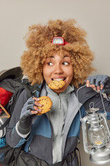Indoor shot of female tourist bites cookie holds lantern looks away wears windbreaker and gloves carries backpack looks away isolated over grey background. Camping time and recreation concept