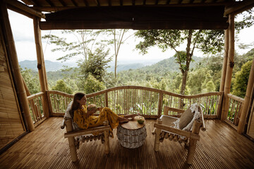 Young  pretty tourist girl sit on veranda with mountains view and drink coconut. Enjoy vacation on Bali island