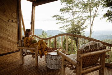 Young  pretty tourist girl sit on terrace of wooden eco house with mountains view and drink...