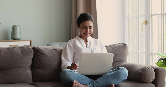 Smiling young Indian woman sit cross legged on cozy sofa hold notebook computer on laps chat in messenger think before write answer. Happy female student relax at home use entertainment app on laptop