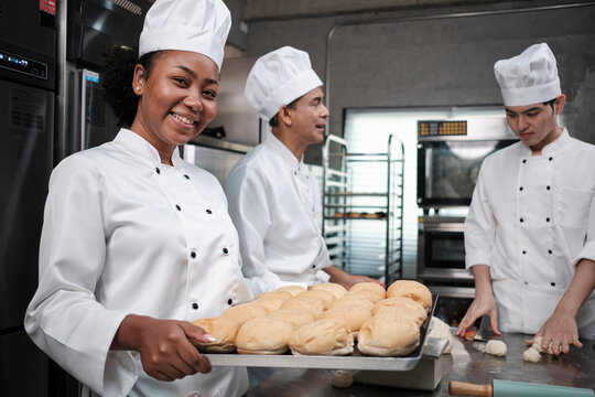 Portrait of African American female chef in white cooking uniform looking at camera with cheerful smile and proud with tray of bread in kitchen, pastry foods professional and fresh bakery occupation.