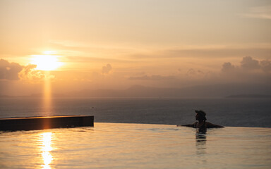 Silhouette of a young woman relaxing in infinity swimming pool and looking at a beautiful sunset and the sea view