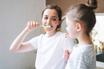Young mother woman with long hair with little tween girl daughter in pajamas brushing their teeth...