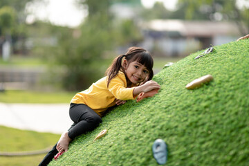 Cute Asian girl having fun trying to climb on artificial boulders at schoolyard playground, Little...