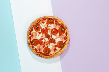 Italian pizza with ham, pepperoni and jalapeno on coloured background. Meat pizza with salami and chilli pepper in minimal style on blue color. American pizza delivery concept with color backdrop