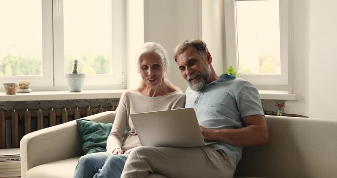 Excited elderly family couple sit on sofa before laptop feel surprised receiving discount proposition read news about great sale in email. Overjoyed senior adult spouses get prize at lottery giveaway