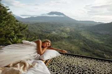 Young woman covering a white blanket sit in hammock  at the view of nature of mountains with green...