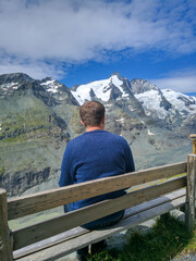 Fototapeta na wymiar Grossglockner, Austria - August 17, 2019: Grossglockner high alpine road against the blue sky. Man sits on a wooden bench and admires the peaks of mountain peaks. Vertical