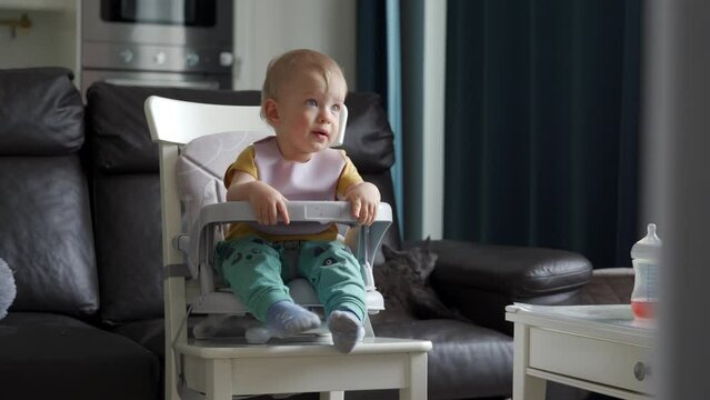 Cute kid sitting in booster seat with feeding tray fixed on top of dining chair, one year old baby sitting in front of tv. High quality 4k footage