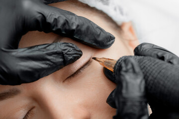 Master makes permanent tattoo makeup on eyelashes of eyes of young woman in beauty salon - obrazy, fototapety, plakaty
