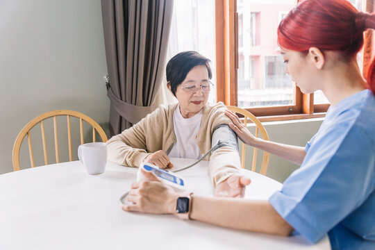 Nurse measuring blood pressure by using automatic blood pressure monitor on senior Asian woman with many medicine bottles of the left side. Caregiver visit. Home health care and nursing home concept.