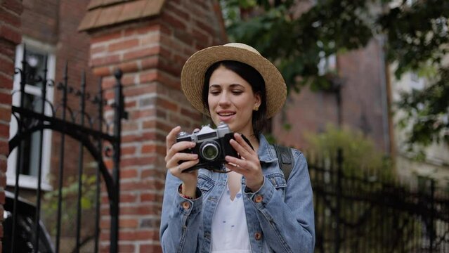 Beautiful young woman in hat taking pictures in the old vintage camera. Sunny day, Outdoors. Happy female walking outdoor in casual denim jacket. Tourist smiling photographer girl make photo.