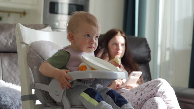 Cute baby boy in booster seat eating an apple, one year old toddler with mother sitting in front of tv, child watching cartoon, mom using mobile phone. High quality 4k footage