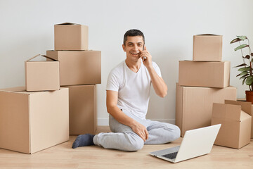 Indoor shot of Caucasian man sitting on floor with laptop before to leave apartment, guy talking on mobile phone among cardboard boxes ordering delivery, looking at camera.