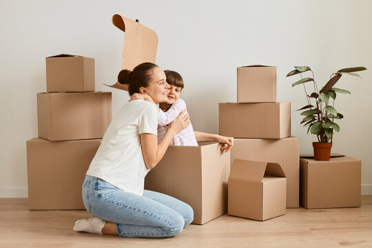 Image of little girl with her mother sitting in handmade boat from cardboard boxes and hugging, happy child playing with mommy during moving in a new flat.