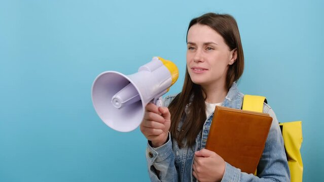 Portrait of promoter girl teen student 20s wears denim jacket and yellow backpack hold books scream in megaphone announces discounts sale Hurry up, posing isolated over blue color background in studio