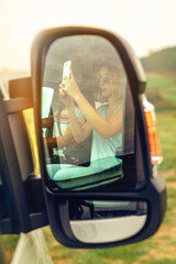 Reflection in a camper van rear-view mirror of a woman taking a picture with her mobile while her...