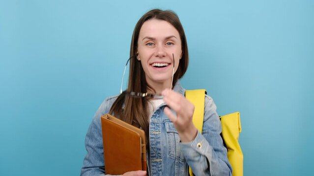 Portrait of girl teen student wears denim jacket and yellow backpack hold books put off glasses say wow, posing isolated on blue color background in studio. Education in university college concept