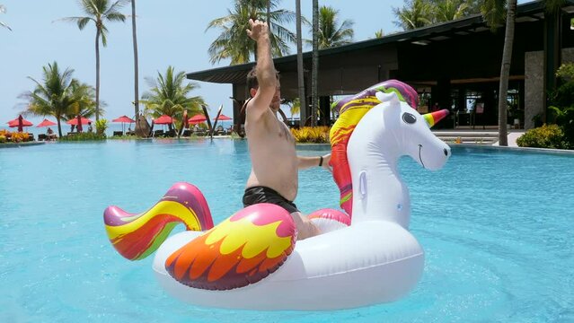 Funny video young man falling in pool in luxury hotel, resort. Happy man have fun ride dance on giant inflatable unicorn in swimming pool, splashing, enjoy holidays or vacations on sunny day in summer