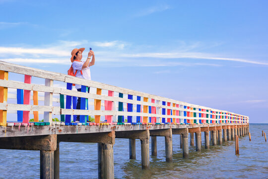 Perspective side view of Asian female tourist photographing with smartphone on colorful wooden bridge at sea viewpoint against cloud on blue sky background 