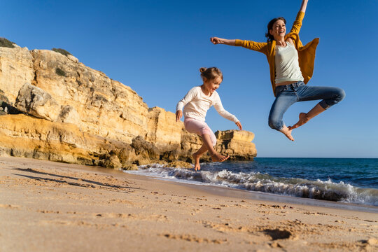 Happy mother with daughter jumping at beach on sunny day