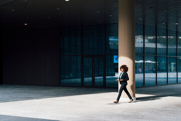 Businesswoman holding tablet PC walking outside office building on sunny day