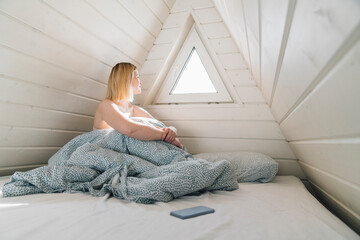 Woman looking out of window sitting on bed in attic