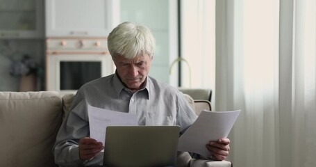 Older serious thoughtful man sit on couch at home read papers, working check budget, personal finances, pay mortgage bills through electronic bank app on laptop, consider paper correspondence concept