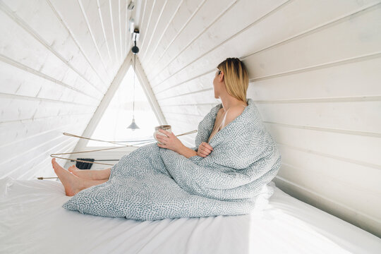 Woman covered in blanket sitting on bed in attic at home