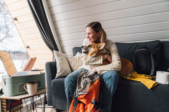 Happy woman with backpack embracing dog sitting on sofa at home