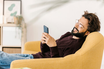 Happy man using smart phone sitting on sofa at home