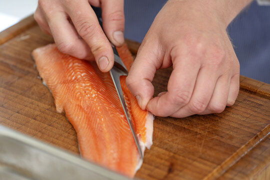 Hands of a cook remove the belly flap with bones of a fresh raw char fish with a filleting knife on a cutting board, selected focus