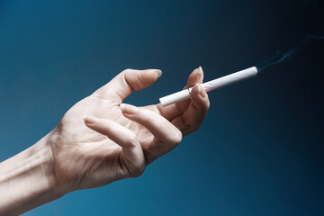 World No Tabacco day. A pale, crooked female hand, close-up, holding a smoking cigarette. Dark blue background. The concept of nicotine addiction