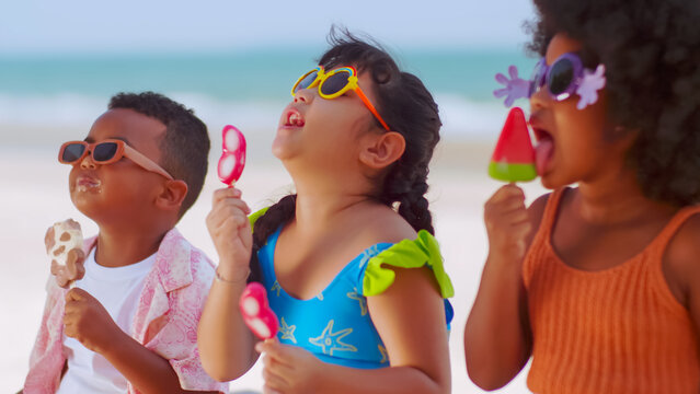 Group Of Children Eating Ice Cream While Having A Picnic On The Beach During Summer Vacation.