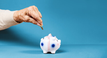 Business banner. Wrinkled hand of an elderly woman puts a euro coin in a piggy bank. Blue...
