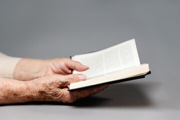 Naklejka premium Wrinkled hands of a senior woman are holding a book with sacred religious texts. Gray background. The concept of religion and prayer