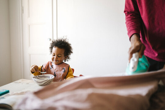 Girl eating from bowl while father ironing clothes