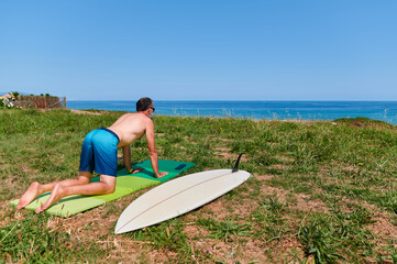 Back view of mature male surfer kneeing on mat on seashore and stretching lower back while preparing for surfing on sunny day in summer