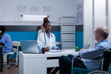 Healthcare clinic medic expert conversating with sick senior patient in wheelchair about illness symptoms and treatment schedule. Hospital doctor talking with elder person with disability about